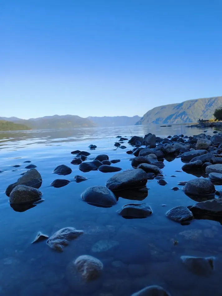 Orilla del Lago Collico con piedras lisas y agua quieta que refleja el cielo, con cerros al fondo
